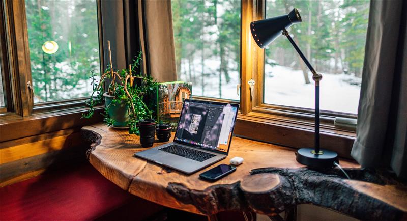 roberto-nickson-1299738-unsplash laptop open on natural wooden desk with cell phone and desk lamp. Facing window and looking out on snowy landscape.