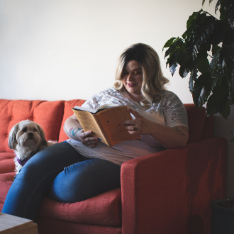 Woman sitting on couch and reading with dog next to her.