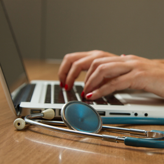 Hands typing on laptop with stethoscope on table.
